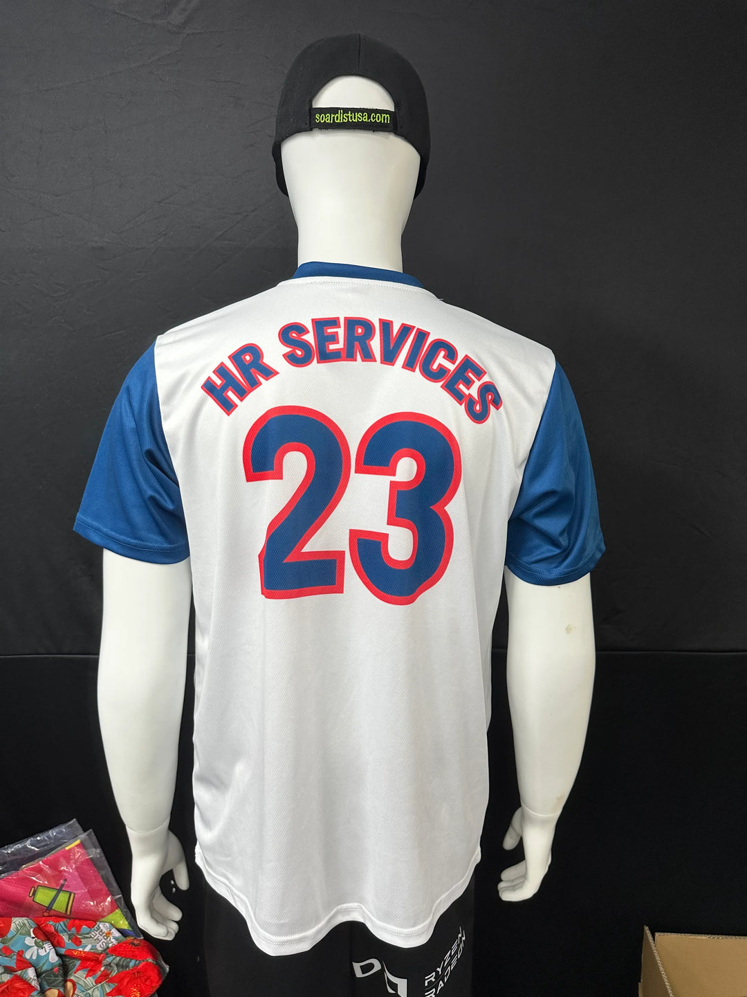 Back view of a white custom athletic jersey with blue sleeves and red 'HR SERVICES 23' text.