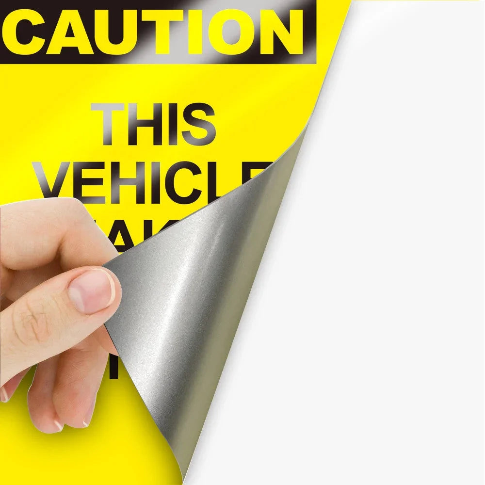 A hand peeling back a silver vinyl layer from a yellow caution vehicle banner.
