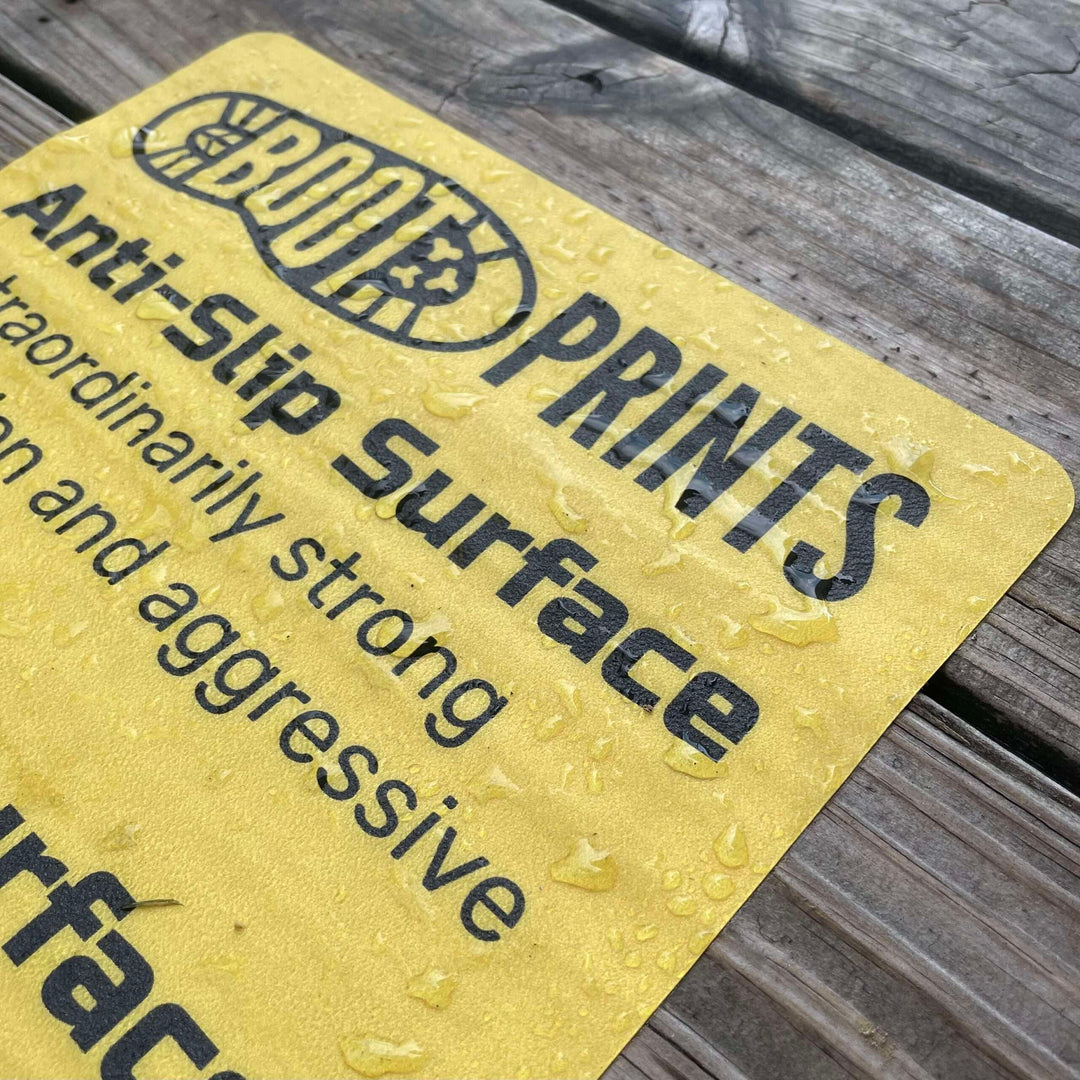 Close-up of a yellow Boot Prints anti-slip surface mat with water droplets on wooden decking.